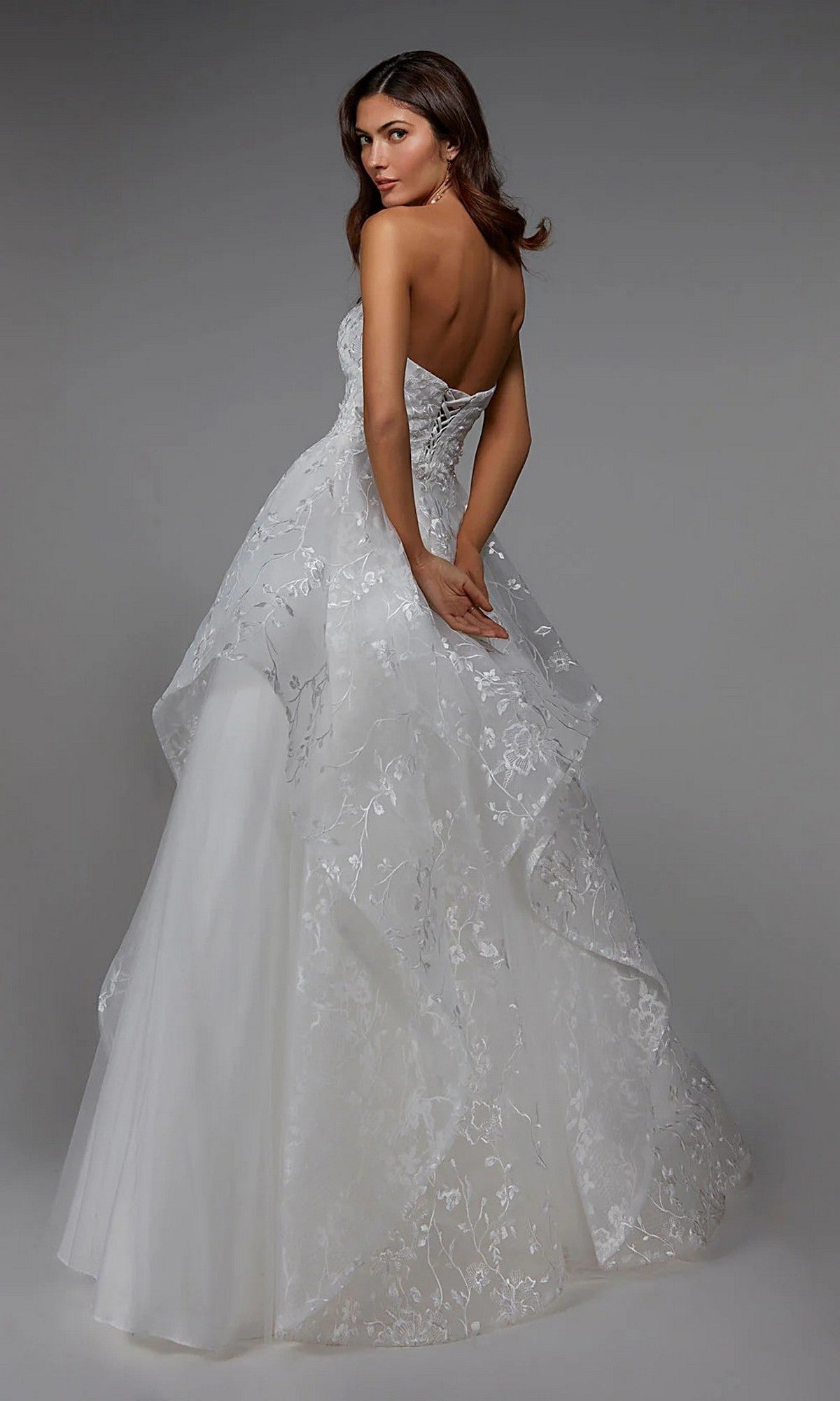 A woman faces away against a plain gray background, wearing the Alyce 7041 long formal dress—a strapless white ball gown featuring a layered, floral-embroidered skirt.